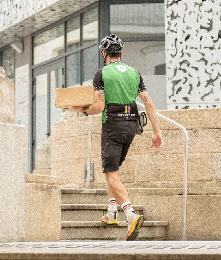 Livreur à vélo en tenue de cyclisme Green Course montant des escaliers en pierre avec un colis devant un bâtiment moderne.
