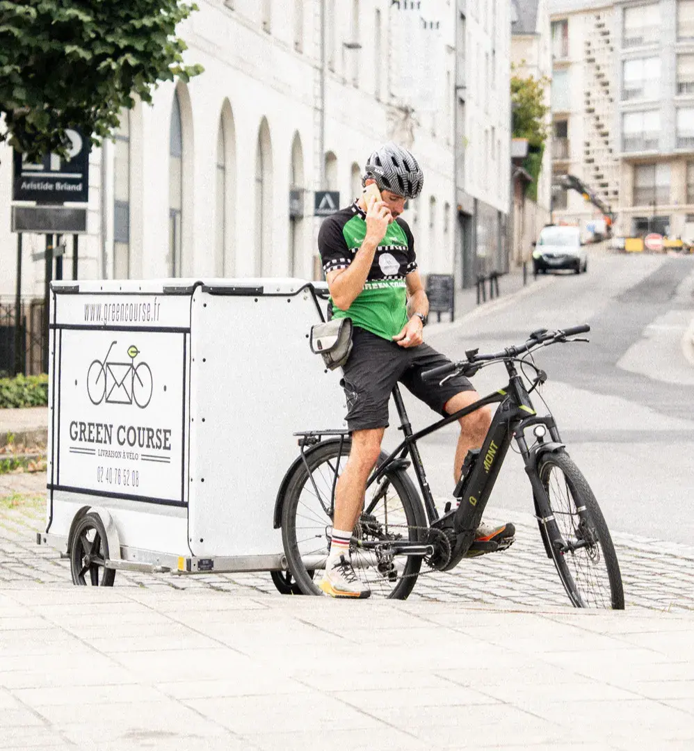 Un livreur à vélo, stationné, avec une remorque caisson de l'entreprise Green Course, est en train de téléphoner.